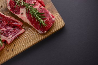 Ahşap kesme tahtasında taze çiğ biftek, siyah arka planda baharatlar, üst manzara..