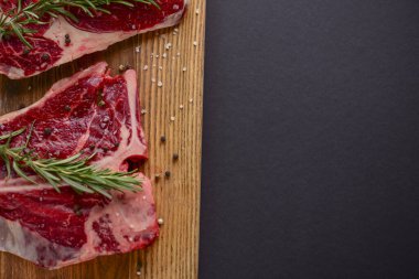 Ahşap kesme tahtasında taze çiğ biftek, siyah arka planda baharatlar, üst manzara..