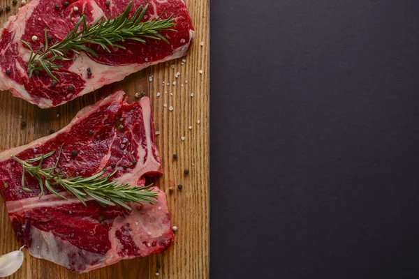 Ahşap kesme tahtasında taze çiğ biftek, siyah arka planda baharatlar, üst manzara..