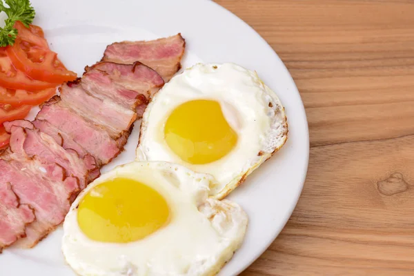 用熏肉、煎蛋和西红柿在淡淡的木桌上的白盘上吃早餐. — 图库照片