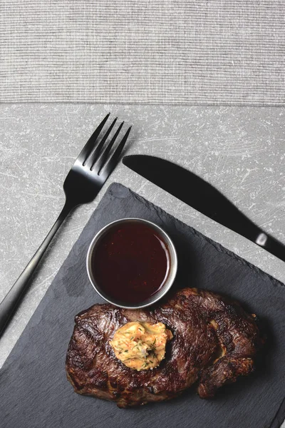 Bife grelhado médio raro com molho em tábua de corte de madeira preta com utensílios. Espaço de cópia, flat lay . — Fotografia de Stock