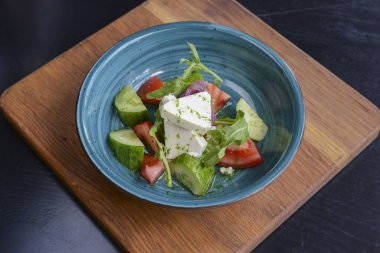 One plate of vegetable salad with feta cheese, cucumbers, tomatoes, and broccoli served in bowl on rustic wooden board.