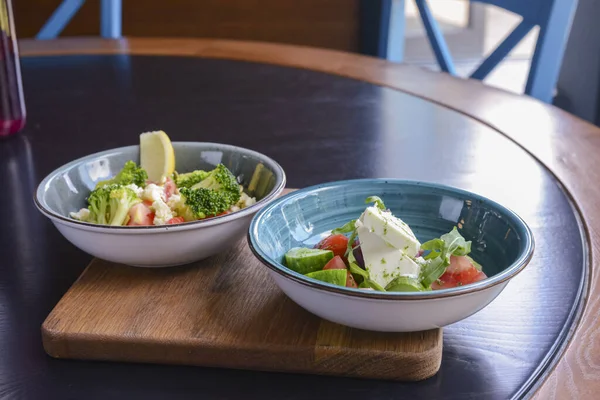 Two plates of vegetable salad with feta cheese, cucumbers, tomatoes, and broccoli served in bowl on rustic wooden board.