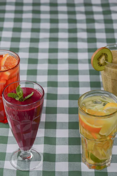 Cztery tropikalne napoje mieszane, jagody, pomarańczowe i kiwi koktajle owocowe serwowane na zielonym obrusie w kratę. — Zdjęcie stockowe