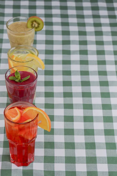 Three tropical mixed drinks, berries, orange and kiwi fruit cocktails served on green plaid tablecloth. — Stock Photo, Image