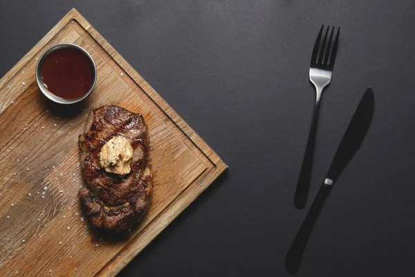 Vue du dessus du barbecue grillé steak de viande fumée avec sauce sur une planche de bois foncé sur fond noir, pose plate, espace de copie — Photo