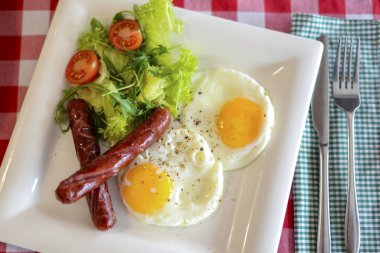 Salatalı kahvaltı, kızarmış yumurta ve kırmızı masa örtüsünün üzerinde beyaz tabakta servis edilen sosisler..