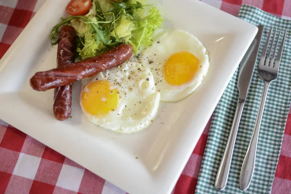 Śniadanie z sałatką, smażone jajko i kiełbaski serwowane w białym talerzu na czerwonym obrusie płyty. — Zdjęcie stockowe