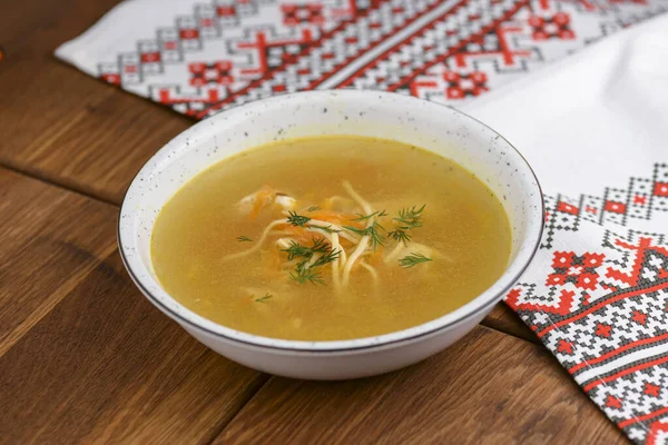 Sup ayam dengan mie dan sayuran disajikan dalam mangkuk putih di atas meja kayu berkarat dengan taplak meja tradisional . — Stok Foto