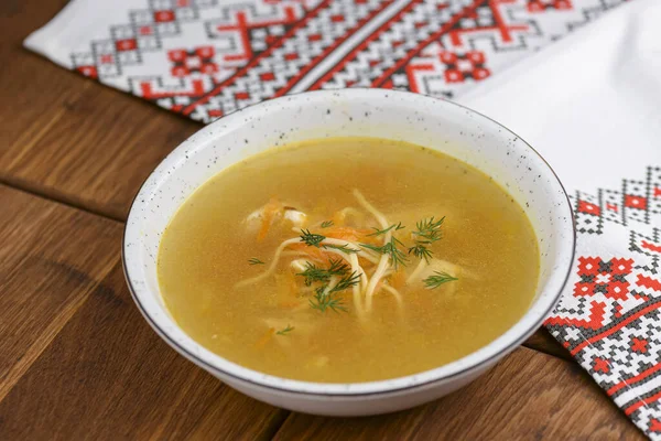 Sup ayam dengan mie dan sayuran disajikan dalam mangkuk putih di atas meja kayu berkarat dengan taplak meja tradisional . — Stok Foto