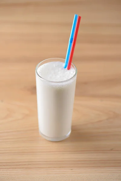 Waniliowy koktajl mleczny w wysokiej galessie z dwiema plastikowymi słomkami na lekkim, drewnianym stole. — Zdjęcie stockowe