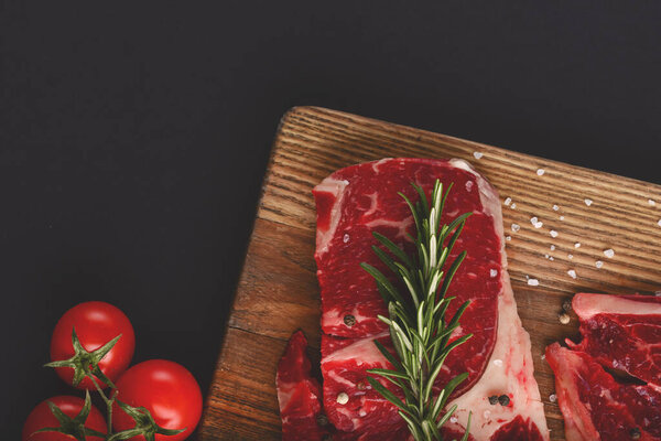 Fresh raw beef steak on wooden cutting board over black background with spices, top view.