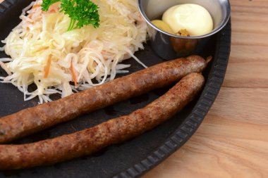 Ekşi lahana salatası ve sosla servis edilen ızgara sosisler, açık sade ahşap arka planda siyah bir tabakta..