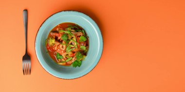 Parlak turuncu arka planda izole edilmiş bir kasede servis edilen sebzeli et salatası ve taze yeşillik..