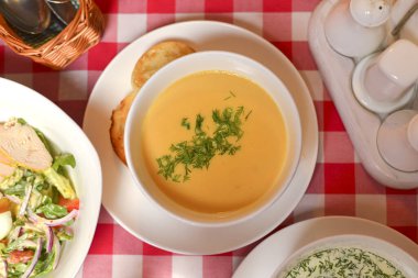 Beyaz kasede balkabağı çorbası. Tahta masa, kırmızı ekose masa örtüsü, bulanık arka plan. Restoranda akşam yemeği.