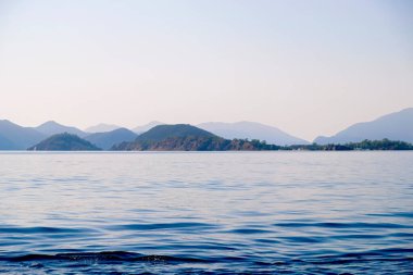 Huzurlu deniz manzarası. Sakin denizin üzerindeki güzel mavi gökyüzü ve güneş ışığı yansıması. Deniz dalgaları. Ufuktaki dağlar.