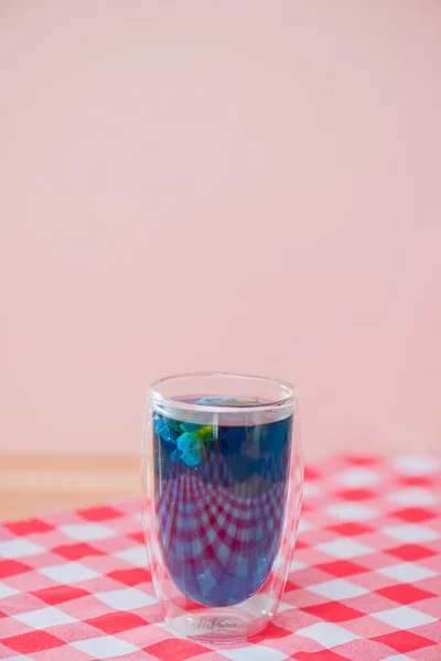 Butterfly pea blue tea in a glass on a plaid red teblecloth. Double wall glass. Copy space banner.