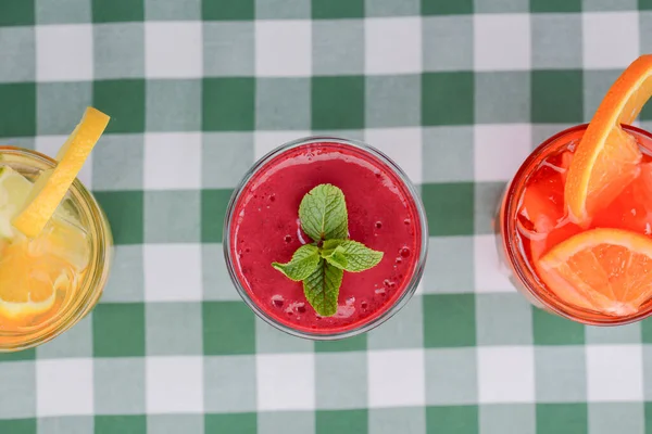 Three tropical mixed drinks, berries, orange and kiwi fruit cocktails served on green plaid tablecloth. — Zdjęcie stockowe