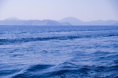 Huzurlu deniz manzarası. Sakin denizin üzerindeki güzel mavi gökyüzü ve güneş ışığı yansıması. Deniz dalgaları. Ufuktaki dağlar.