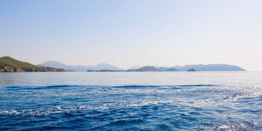 Huzurlu deniz manzarası. Sakin denizin üzerindeki güzel mavi gökyüzü ve güneş ışığı yansıması. Deniz dalgaları. Ufuktaki dağlar.