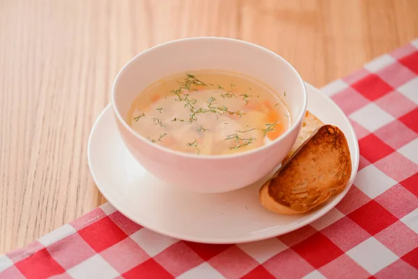 Chicken soup with vegetables and meat served in a white bowl over rustic wooden background with red plaid tablecloth. — Stock Photo, Image