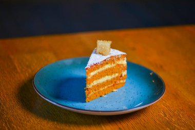 Katmanlı vanilyalı ballı kek, bulanık restoranın arka planında, ahşap bir masada mavi bir tabakta servis ediliyor..