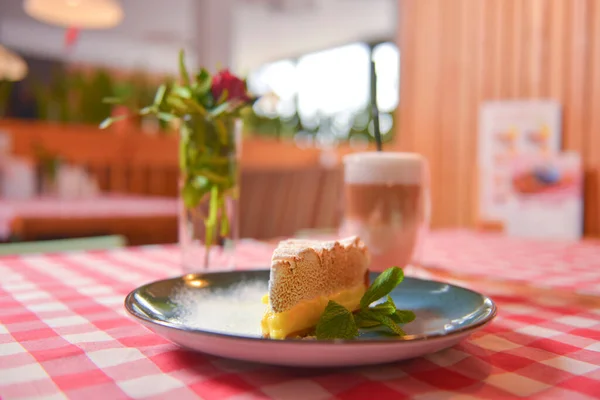 Piece of cream lemon meringue pie served with Latte coffee on a table with red plaid tablecloth.