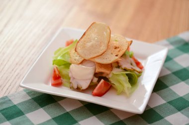 Tavuk ve salatalı geleneksel Sezar Salatası tabağı açık ahşap masa ve yeşil ekose masa örtüsü..