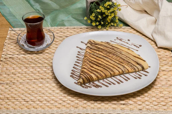畳んだクレープ、布ポーチ、黄色の花とトルコのお茶と一緒に出される白い皿にチョコレート ソースとロシアのブリニ — ストック写真