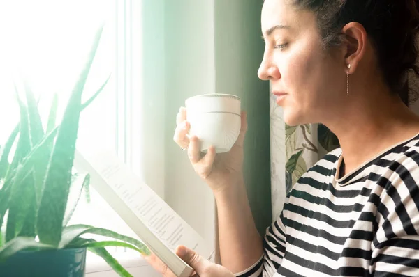 Adult Caucasian woman sitting near window at home relaxing in her living room reading book and drinking coffee or tea