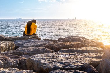 Deniz kenarındaki bir kayanın üzerinde oturan sarı gömlekli bir adamın arka görüntüsü. Boş zaman ve rahatlatıcı konsept.