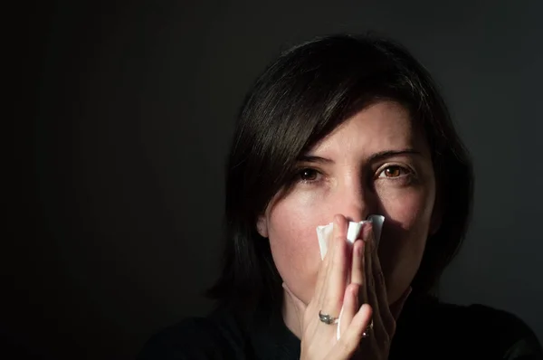 Portrait of a sick brunette woman having flu and high fever, sneezing into tissue on a dark background. Medical and health concept.