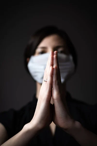 Closeup hands of praying adult woman wearing hygienic mask to prevent infection, airborne respiratory illness such as flu, 2019-nCoV on dark background.