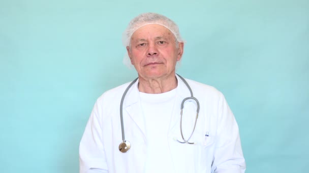 マスクをつけている高齢者の医師は 薄い青の背景に白いコートと青い手袋をしています コロナウイルスに対する保護具を身に着けている古い医師Sars Cov 2の普及と感染症 — ストック動画
