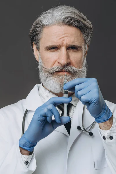 Médico de cabelo cinza sênior segurando uma seringa e olhando para a câmera — Fotografia de Stock