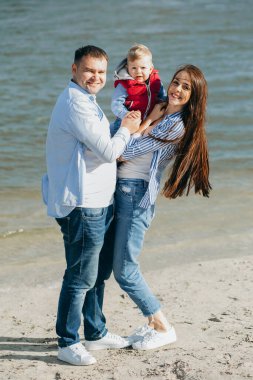 Mutlu genç aile deniz kenarında yeşil doğada birlikte vakit geçiriyor. Ebeveynler dışarıda çocuklarla oynuyor. Aile gün batımında yürüyor, sarılıyor ve eğleniyor.