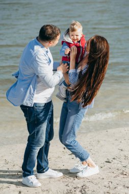 Mutlu genç aile deniz kenarında yeşil doğada birlikte vakit geçiriyor. Ebeveynler dışarıda çocuklarla oynuyor. Aile gün batımında yürüyor, sarılıyor ve eğleniyor.