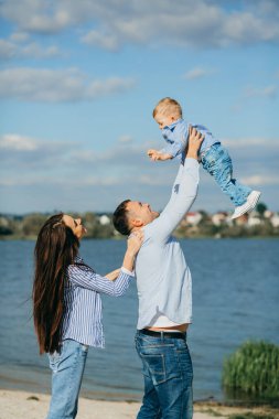 Mutlu genç aile deniz kenarında yeşil doğada birlikte vakit geçiriyor. Ebeveynler dışarıda çocuklarla oynuyor. Aile gün batımında yürüyor, sarılıyor ve eğleniyor.