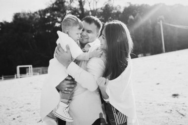 Mutlu genç aile deniz kenarında yeşil doğada birlikte vakit geçiriyor. Ebeveynler dışarıda çocuklarla oynuyor. Aile gün batımında yürüyor, sarılıyor ve eğleniyor.