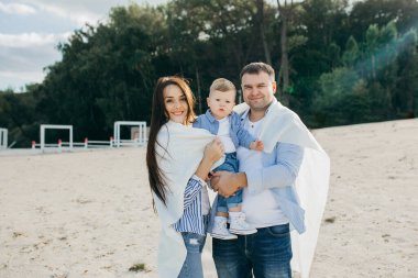 Mutlu genç aile deniz kenarında yeşil doğada birlikte vakit geçiriyor. Ebeveynler dışarıda çocuklarla oynuyor. Aile gün batımında yürüyor, sarılıyor ve eğleniyor.