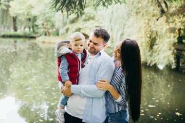 Mutlu genç aile deniz kenarında yeşil doğada birlikte vakit geçiriyor. Ebeveynler dışarıda çocuklarla oynuyor. Aile gün batımında yürüyor, sarılıyor ve eğleniyor.