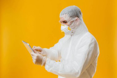 Tehlikeli madde giysisi ve solunum maskesi takmış epidemiyoloji uzmanı sarı arka planda dijital tablet tutuyor.