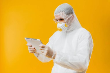 Tehlikeli madde giysisi ve solunum maskesi takmış epidemiyoloji uzmanı sarı arka planda dijital tablet tutuyor.