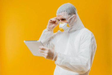 Tehlikeli madde giysisi ve solunum maskesi takmış epidemiyoloji uzmanı sarı arka planda dijital tablet tutuyor.