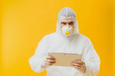 Tehlikeli madde giysisi ve solunum maskesi takmış epidemiyoloji uzmanı sarı arka planda dijital tablet tutuyor.