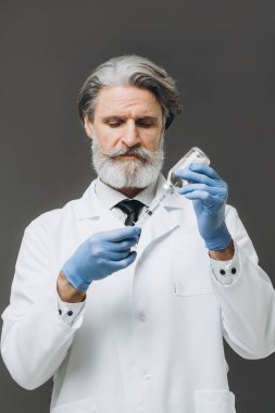 Beyaz önlük, eldiven ve maske giymiş gri saçlı doktor aşıyı enjekte etmeye hazırlanıyor. Gri renkte izole edilmiş.