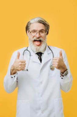 Sarı arka plan üzerinde steteskop takmış yaşlı gri saçlı doktor mutlu ve komik yüzlü parmaklarını kameraya doğrultuyor. İyi enerji ve titreşimler.