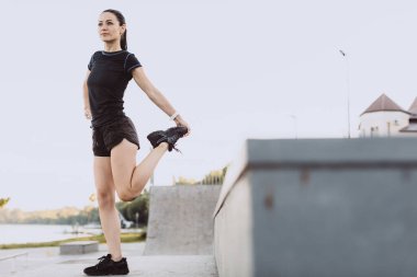 Siyah giyinmiş atletik kız, sabah şehir plajında koşmadan önce ısınıyor.