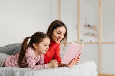 Mutlu aile annesi ve çocuğu kitap okuyor, gülümsüyor anne, bebek bakıcısı tatlı anaokulu kızına komik masallar anlatıyor, birlikte gülüyor ve yatmadan önce masallar anlatıyor.