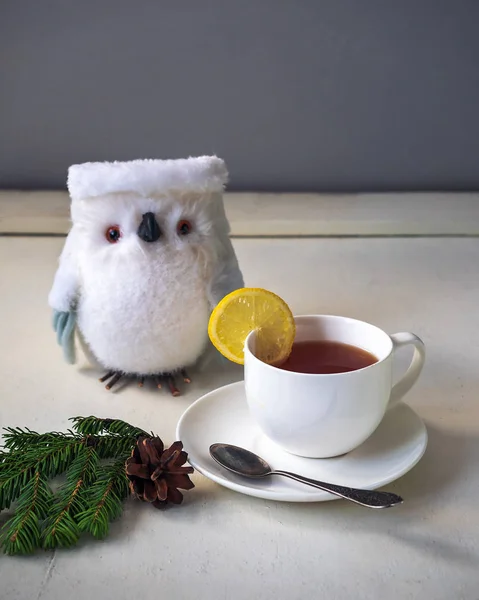 Té con limón en una taza blanca sobre un platillo, servido sobre una mesa blanca con un búho decorativo . — Foto de Stock